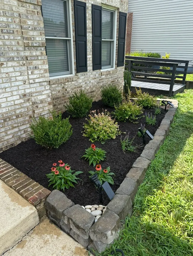 Mulched flower bed with a stacked-stone border and red blooms beside a brick home
